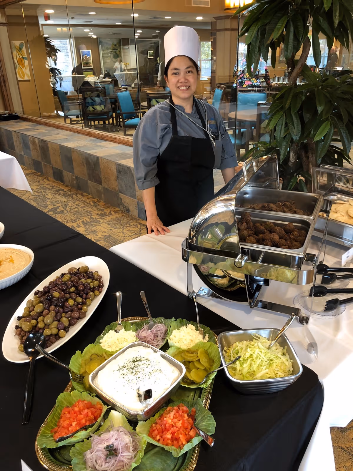 A smiling chef stands behind a buffet table with chafing dishes and bowls of olives, salads, and condiments in a dining room.