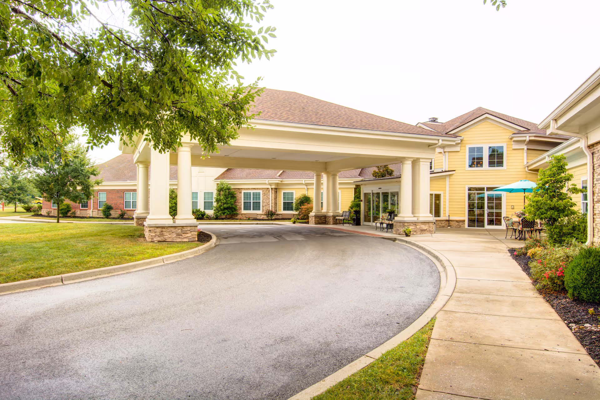 Entrance of a senior living facility with a covered driveway supported by columns, surrounded by well-maintained landscaping and outdoor seating with an umbrella.