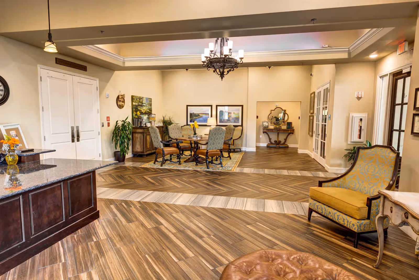 A spacious and well-lit common area in a senior living facility featuring a wooden floor with a patterned design. There is a round table with four upholstered chairs on a rug in the center, a chandelier hanging from the ceiling, and various decorative elements including framed paintings, plants, and a sideboard with a beverage dispenser. To the right, there is a yellow and blue patterned armchair next to a small table, and to the left, a granite countertop with a vase of flowers.