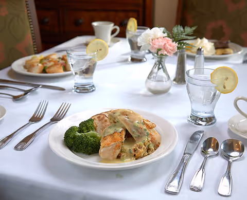 A dining table set with white tablecloth featuring plates of food including chicken with sauce and broccoli, glasses of water with lemon slices, silverware, and small vases with pink and white flowers.