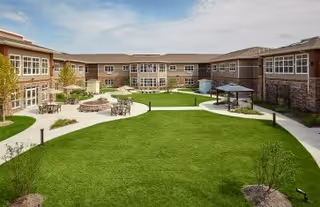 A spacious outdoor courtyard area of a senior living facility with well-maintained green lawns, paved walkways, seating areas with tables and chairs, and a gazebo. The courtyard is surrounded by a two-story building with large windows under a partly cloudy sky.