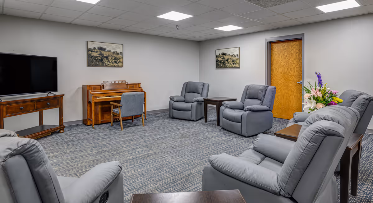 A common area with six gray recliner chairs arranged in a circle on a blue carpeted floor. There is a wooden piano with a chair in front of it against the wall, a flat-screen TV on a wooden stand, two small wooden side tables, a wooden door, two landscape paintings on the walls, and a floral arrangement on a side table.