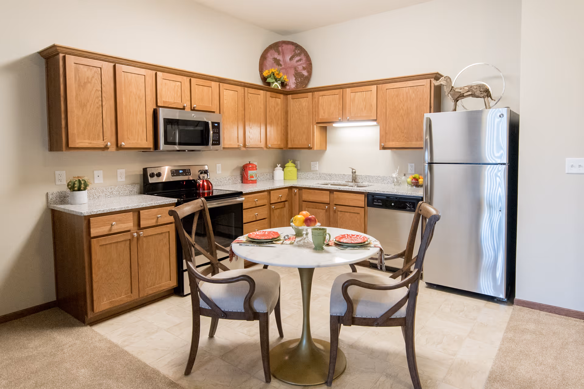 A kitchen area with wooden cabinets, a stainless steel refrigerator, stove, microwave, and dishwasher. In the center, there is a small round dining table set for two with plates, cups, and a bowl of fruit. The floor is a combination of tile and carpet.