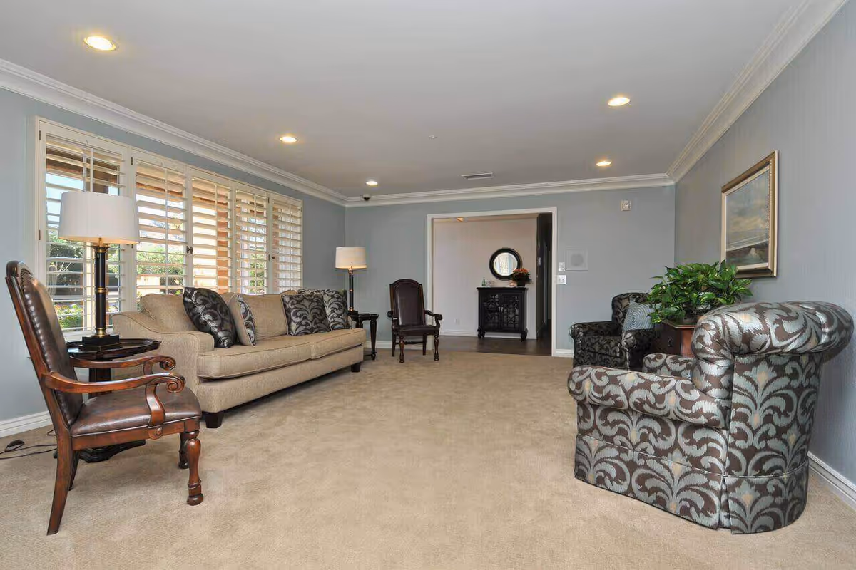 A spacious living room with beige carpet and light blue walls. The room features a beige sofa with patterned cushions, two patterned armchairs, and a wooden chair with leather upholstery. There are two floor lamps with white shades, a large window with white shutters, a framed painting on the wall, and a potted plant on a side table. In the background, there is an open doorway leading to another room with a black cabinet and a round mirror above it.