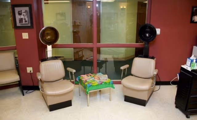 Two beige salon chairs with vintage hair dryers attached, positioned on either side of a small table covered with a green cloth and magazines, set against a maroon wall with a large window and a beige chair to the left.