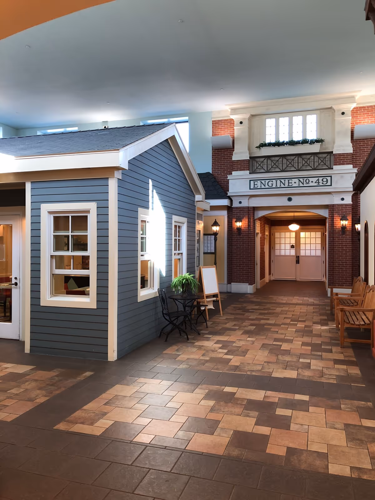 Indoor hallway area designed to look like a small town street with a blue house facade on the left, a brick building facade labeled 'ENGINE No. 49' straight ahead, tiled floor, a small black table with two chairs and a potted plant, and wooden benches on the right.