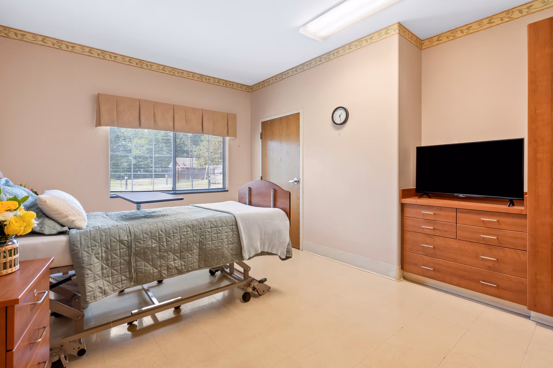 A clean and well-lit patient room in a healthcare facility featuring a hospital bed with green bedding and pillows, a wooden dresser with a flat-screen TV on top, a window with blinds and a beige valance, a wooden door, a wall clock, and a small table attached to the bed. There is a vase with yellow flowers on a bedside table.