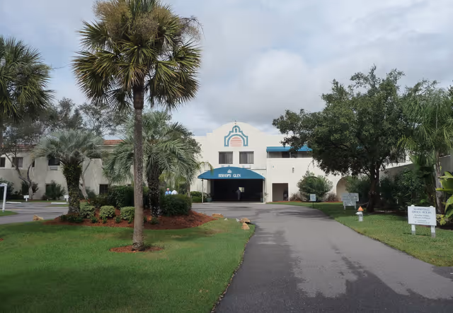 Front exterior view of Bishops Glen Retirement Center showing a driveway leading to the entrance with a blue awning, surrounded by green lawns, palm trees, and other landscaping under a cloudy sky.