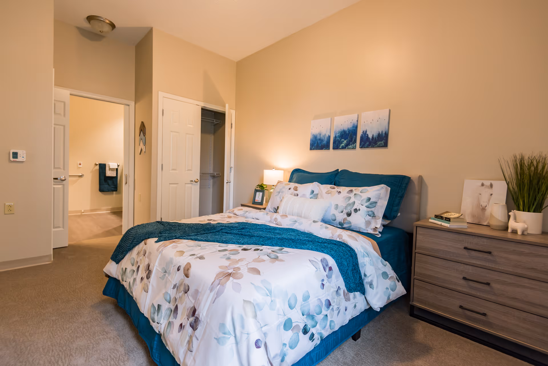 A cozy bedroom in a senior living facility featuring a neatly made bed with floral bedding and teal accents. The room includes a wooden dresser with decorative items and a small lamp on a nightstand. There are three nature-themed paintings on the wall above the bed. An open door reveals a glimpse of an adjacent bathroom with towels hanging on the wall.