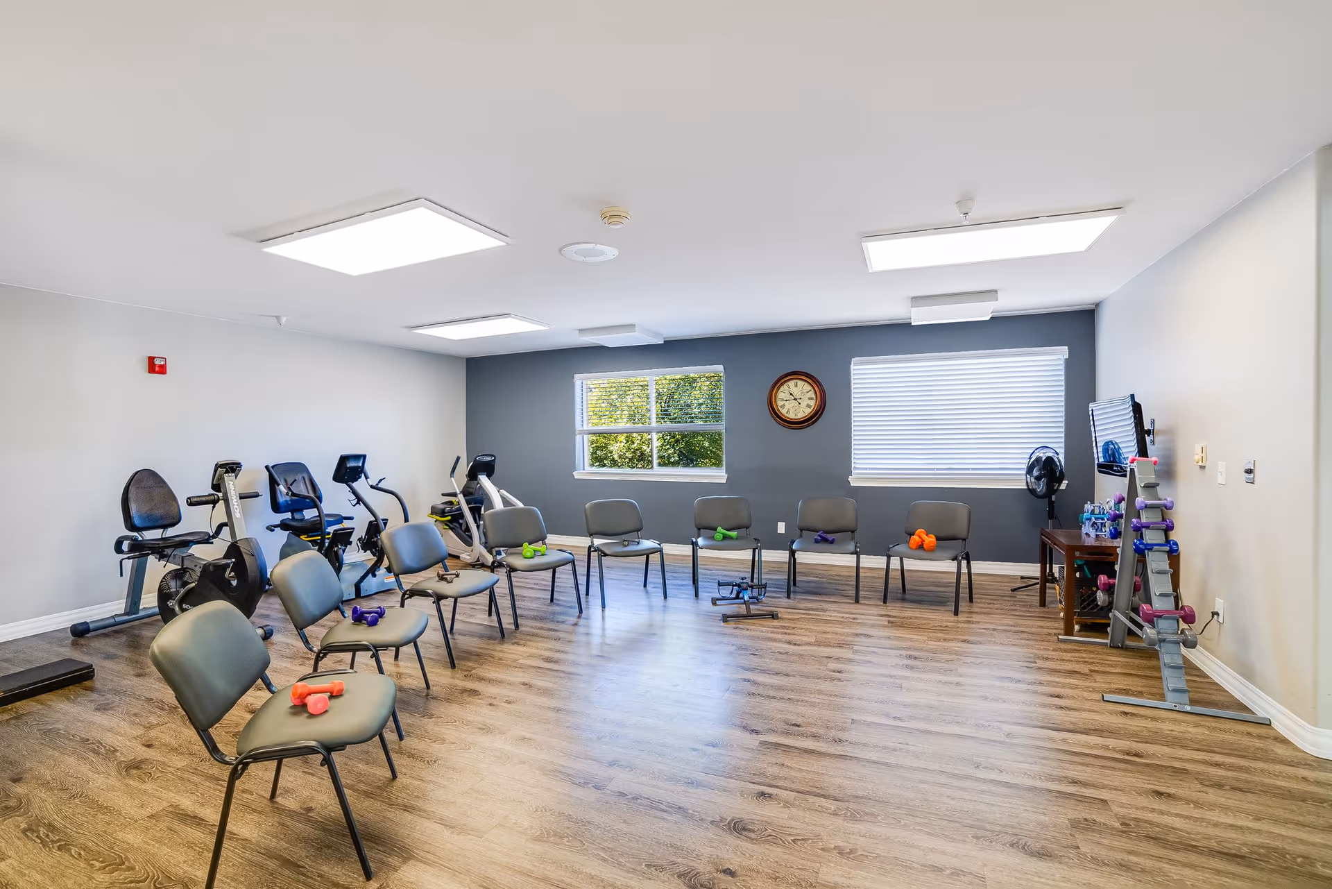 Bright exercise room with chairs arranged in a circle, stationary bikes, hand weights and a wall clock.