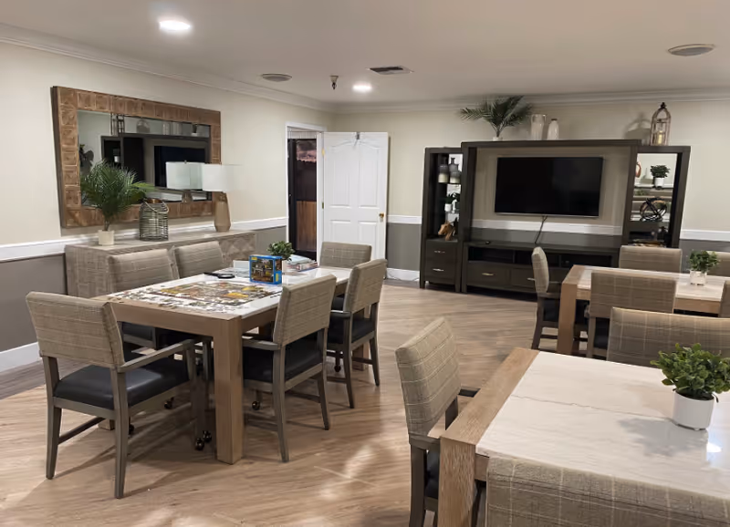 A well-lit common area with multiple tables and chairs arranged for seating. The room features a large mirror on one wall, a TV mounted on a dark wood entertainment unit with shelves and decorative items, and light-colored walls with a chair rail. The floor is wood, and there are small potted plants on the tables.
