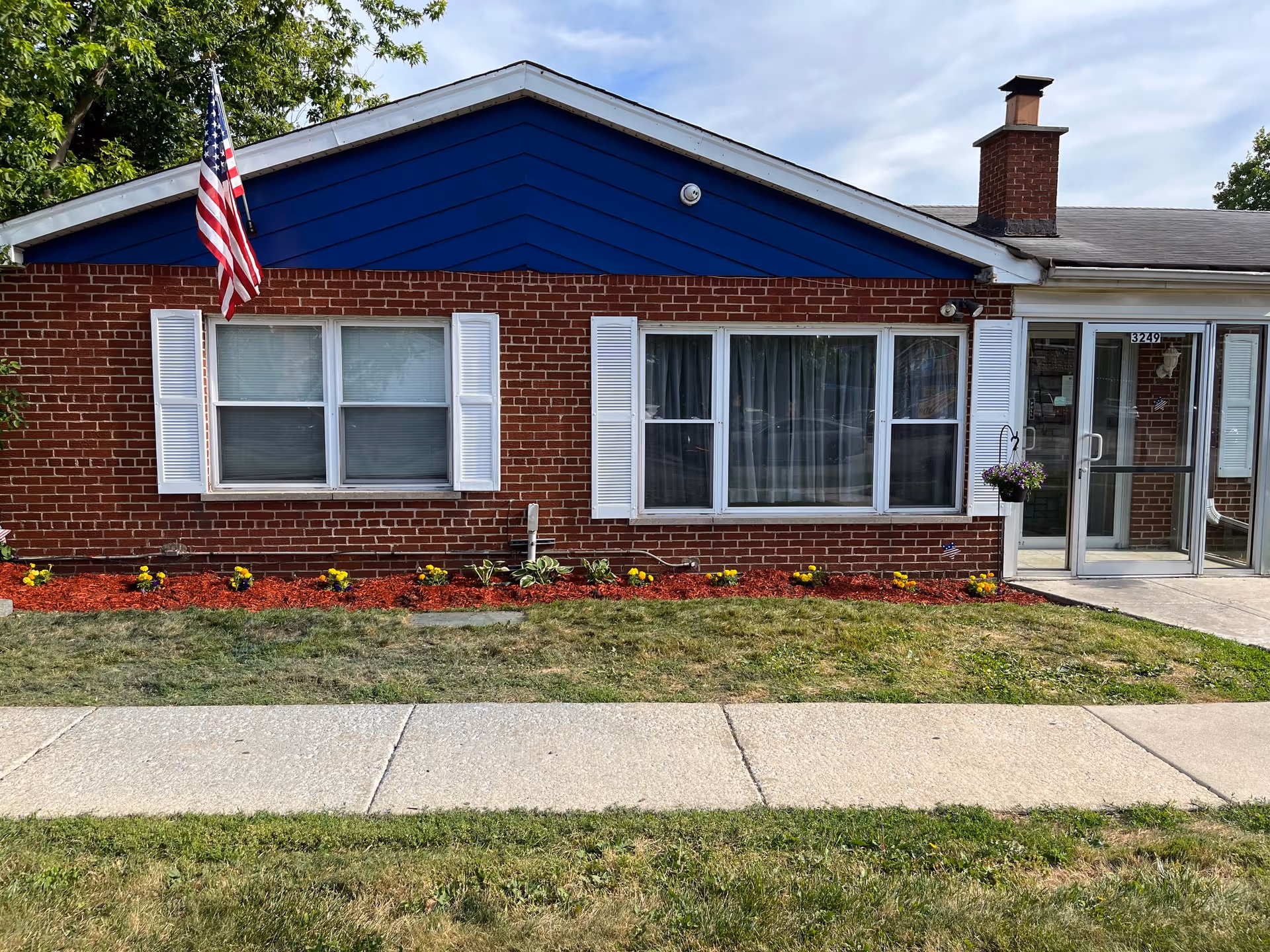 Front exterior of a single-story brick building with white-trimmed windows, an American flag, flowerbeds, and a glass entrance on the right.