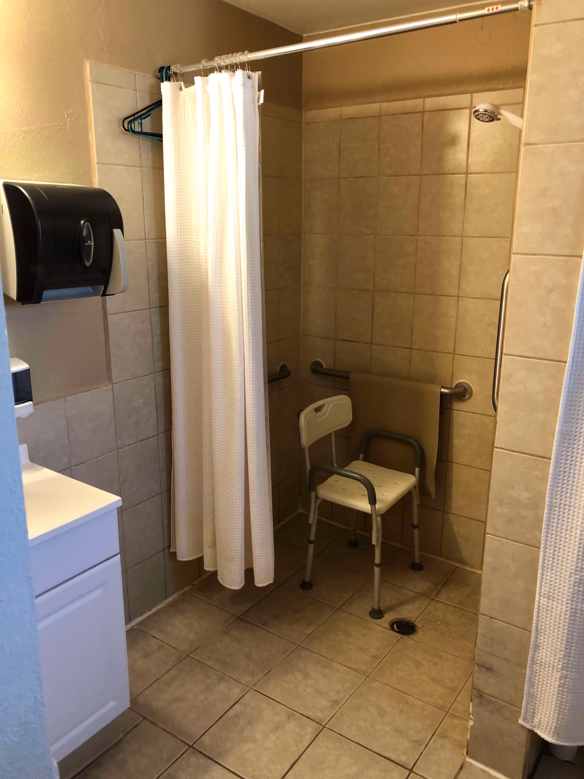A tiled bathroom with a walk-in shower area featuring a white shower chair with armrests, grab bars on the walls, and a beige towel hanging on one of the bars. A white shower curtain is partially drawn, and there is a black paper towel dispenser mounted on the wall next to a white sink cabinet.