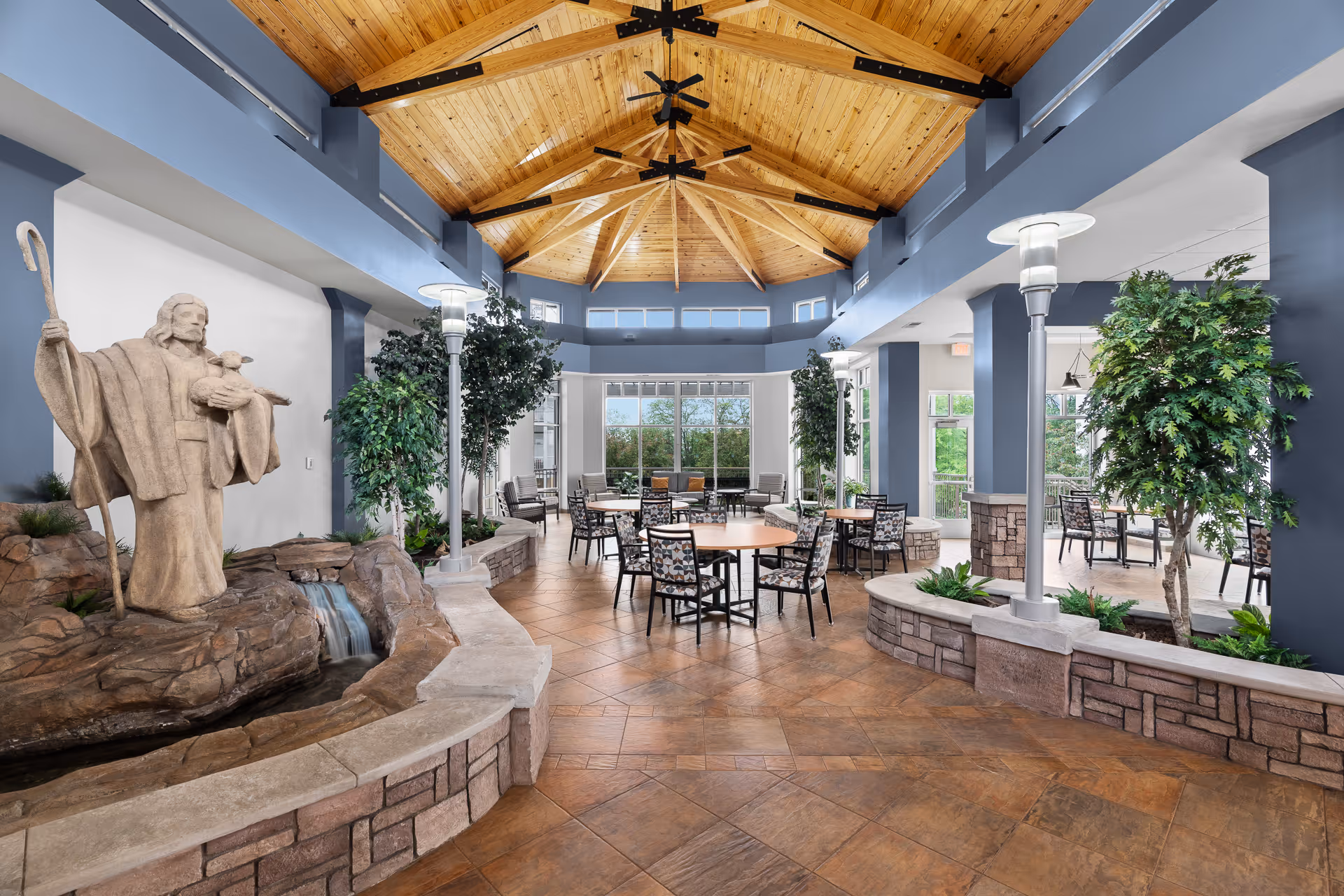 Spacious indoor common area with a high wooden ceiling featuring black metal beams and ceiling fans. The room has large windows allowing natural light to fill the space. Several round tables with patterned chairs are arranged throughout the area. There are tall green plants and stone planters along the sides. A stone statue of a shepherd holding a lamb stands beside a small water feature with rocks and a waterfall on the left side.