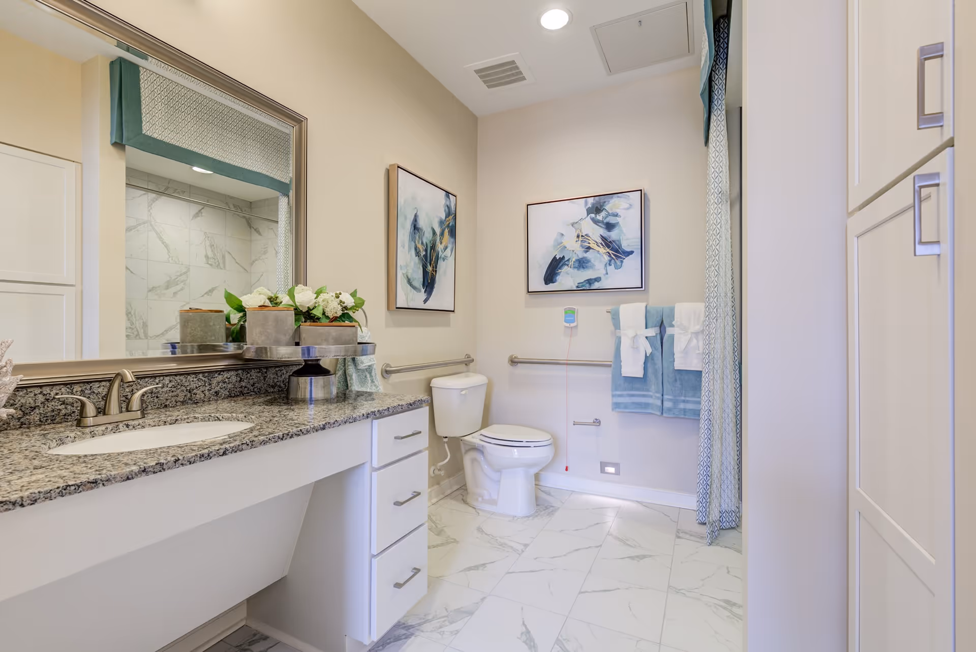 A clean and modern bathroom with a granite countertop sink, a large mirror, a toilet with grab bars on the wall, two blue towels hanging on a towel rack, abstract artwork on the walls, and white cabinets. The floor is tiled with white marble-patterned tiles.