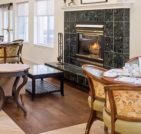 Cozy interior dining area with wooden chairs featuring floral upholstery, a round dining table set with white dishes and glassware, a black tiled fireplace with a fire burning, and large windows with white blinds allowing natural light to fill the room.