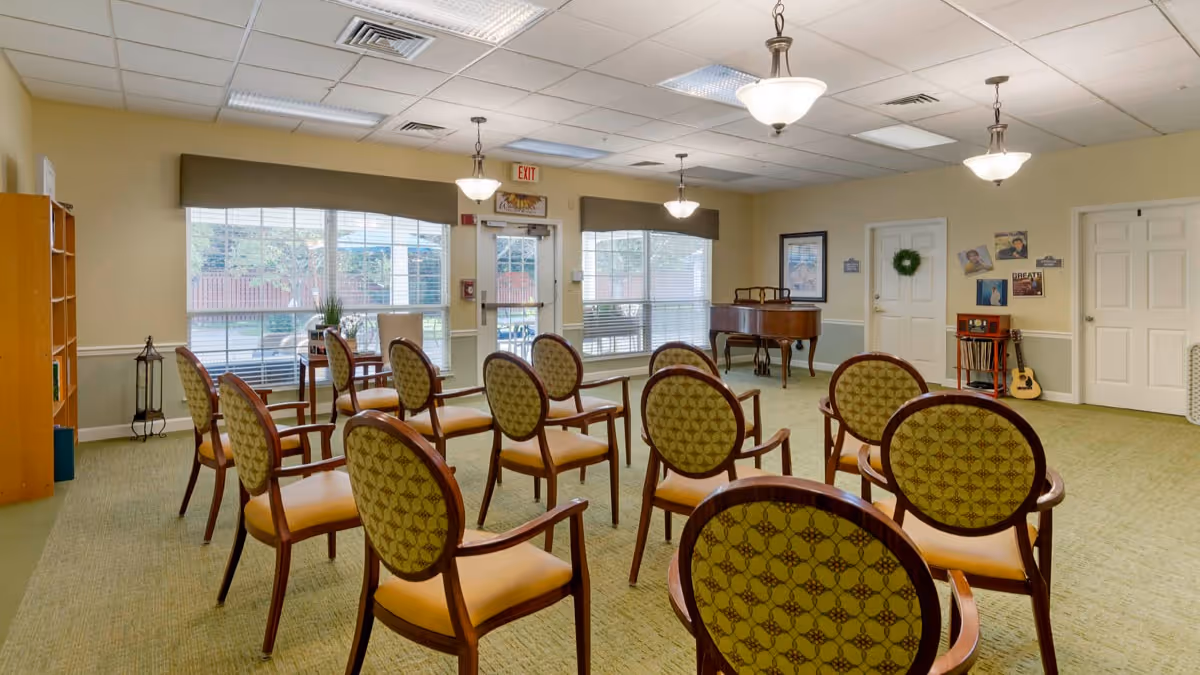 Well-lit activity room with rows of patterned chairs facing a piano and large windows.