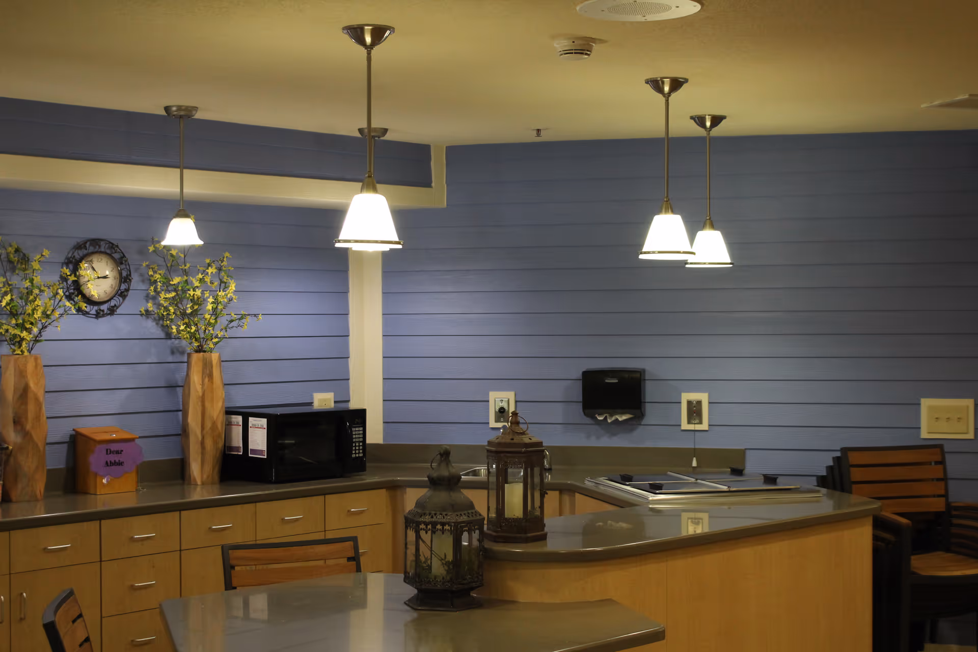 Interior view of a kitchen area with light wood cabinets and a gray countertop. The walls are painted blue with horizontal paneling. There are three pendant lights hanging from the ceiling. On the counter, there is a microwave, two tall vases with yellow flowers, a clock on the wall, a box labeled 'Dear Abbie,' and two decorative lanterns on the island. There are wooden chairs around the island.