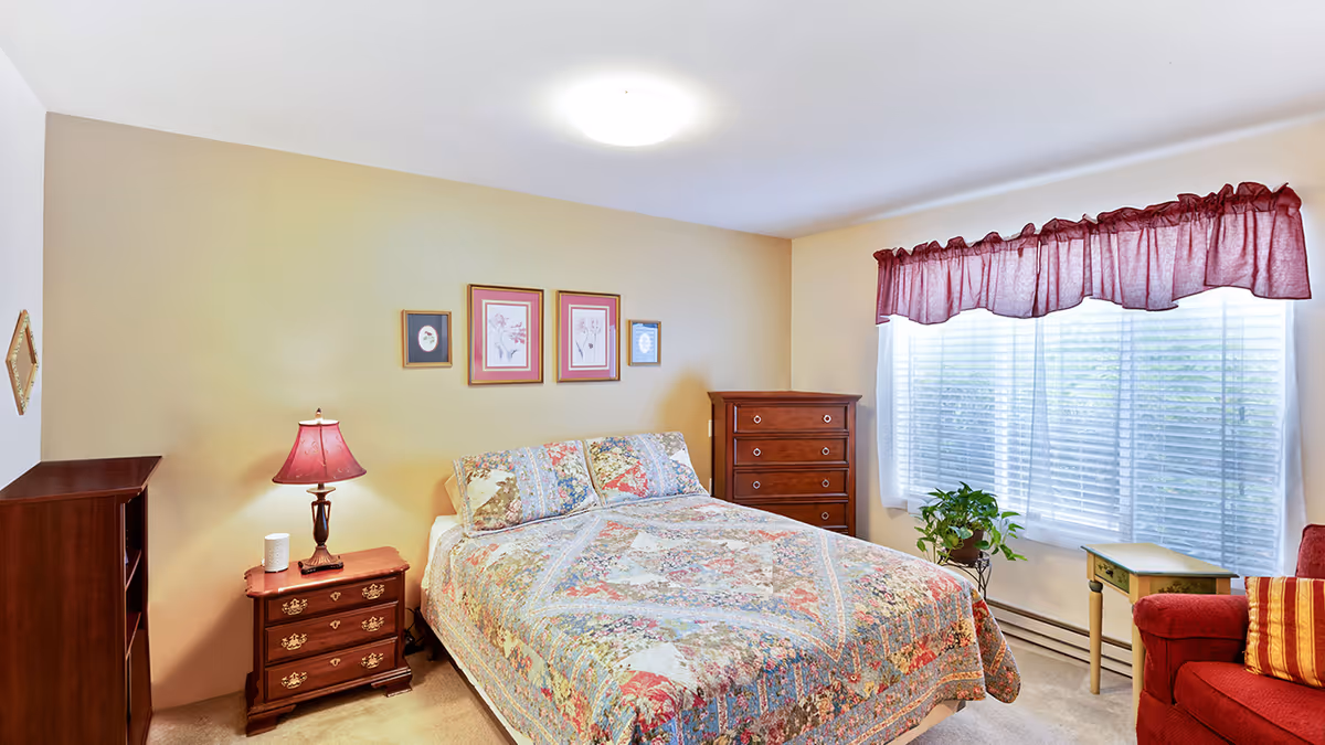 A cozy bedroom with a floral patterned bedspread on a double bed, a wooden nightstand with a red lamp and a small white object, a wooden dresser, a red armchair with a striped pillow, a small green side table, a potted plant near a large window with white blinds and a red valance, and framed floral artwork on the beige walls.