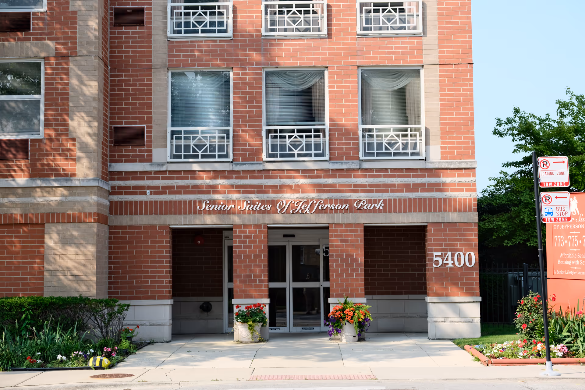Entrance of a brick building with the sign 'Senior Suites of Jefferson Park' above the doorway. The building number 5400 is displayed on the right side of the entrance. There are flower pots with colorful flowers on either side of the entrance and a sidewalk leading up to the door. A signpost with parking and bus stop signs is visible on the right side near a landscaped area with flowers and greenery.