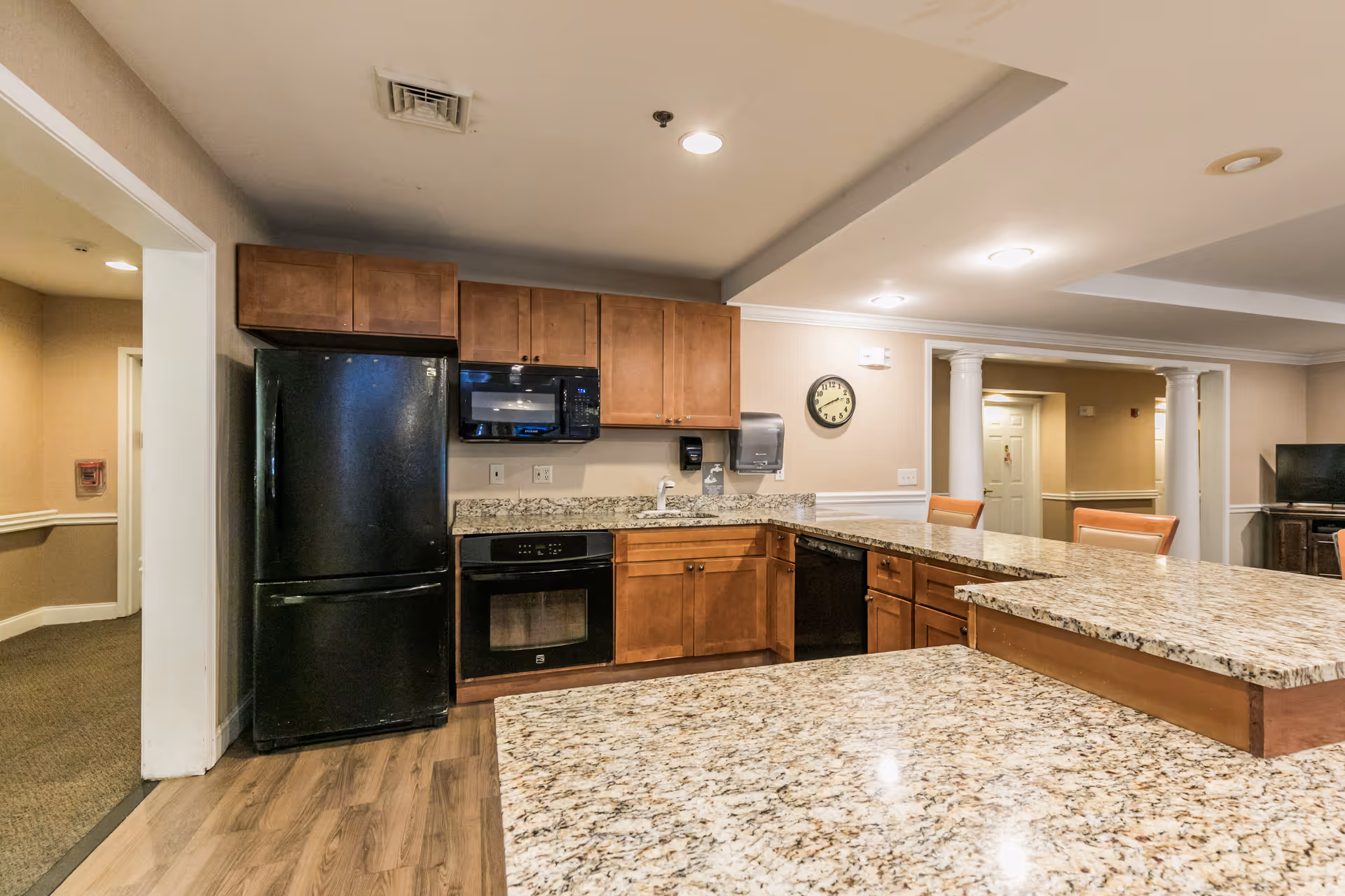 Open kitchen with granite countertops, wooden cabinets, black appliances and bar seating in a senior living facility common area.