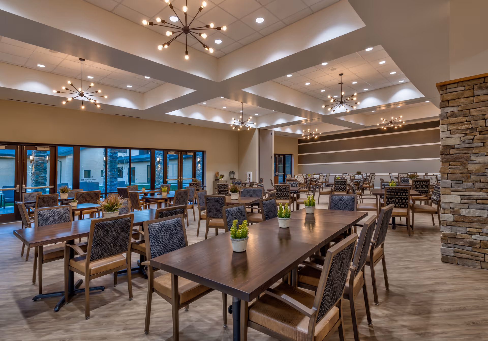 Spacious, well-lit dining room with multiple wooden tables and chairs, modern chandeliers, potted plants on tables, and large windows.