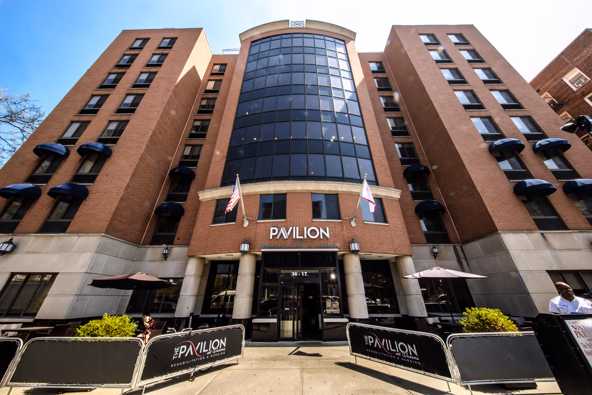 Front exterior view of The Pavilion at Queens for Rehabilitation & Nursing, a multi-story brick building with large glass windows in the center. Two flags are displayed above the entrance, and outdoor seating with umbrellas is visible in front of the building.