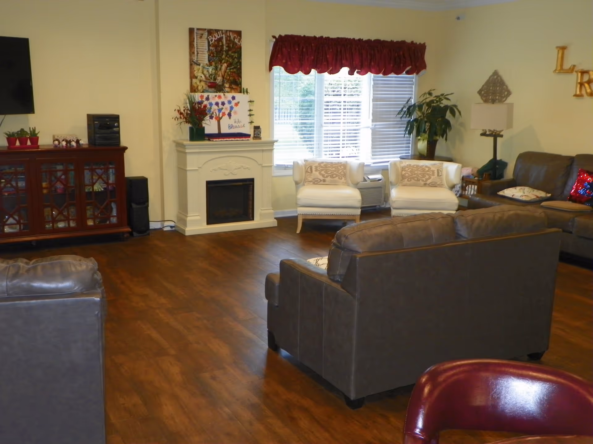 A cozy living room with wooden flooring, featuring a white fireplace with decorative items on top, two white armchairs with patterned cushions near a window with blinds and a red valance, a brown leather sofa and loveseat, a wooden cabinet with glass doors, a flat-screen TV mounted on the wall, and various plants and decorations.