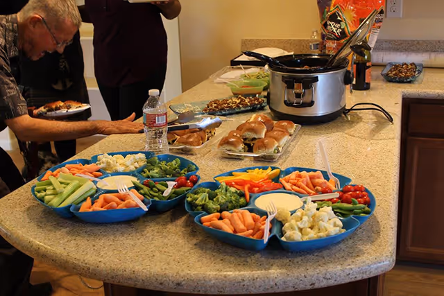 A kitchen counter with trays of fresh vegetables including carrots, celery, broccoli, cauliflower, cherry tomatoes, and dip. There are also plates of sandwiches, a crockpot, a bottle of water, and a bag of chips. Two people are partially visible, one elderly man serving himself food.