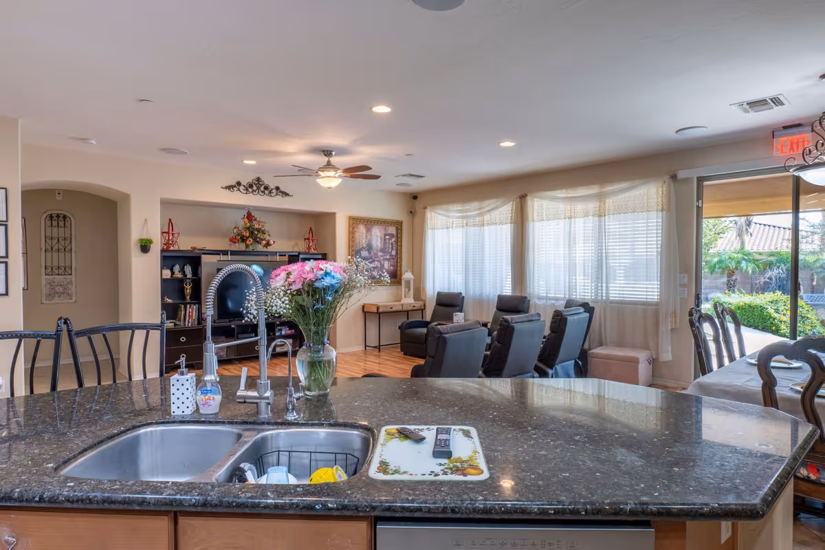 Open-plan senior living interior showing a kitchen island with sink and flowers in the foreground and a seating area with recliners and large windows in the background.