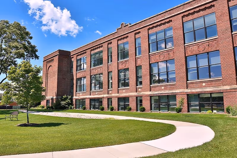 Exterior view of a three-story red brick building with large windows, surrounded by a well-maintained lawn, a curved concrete walkway, a tree, and a bench under a partly cloudy blue sky.