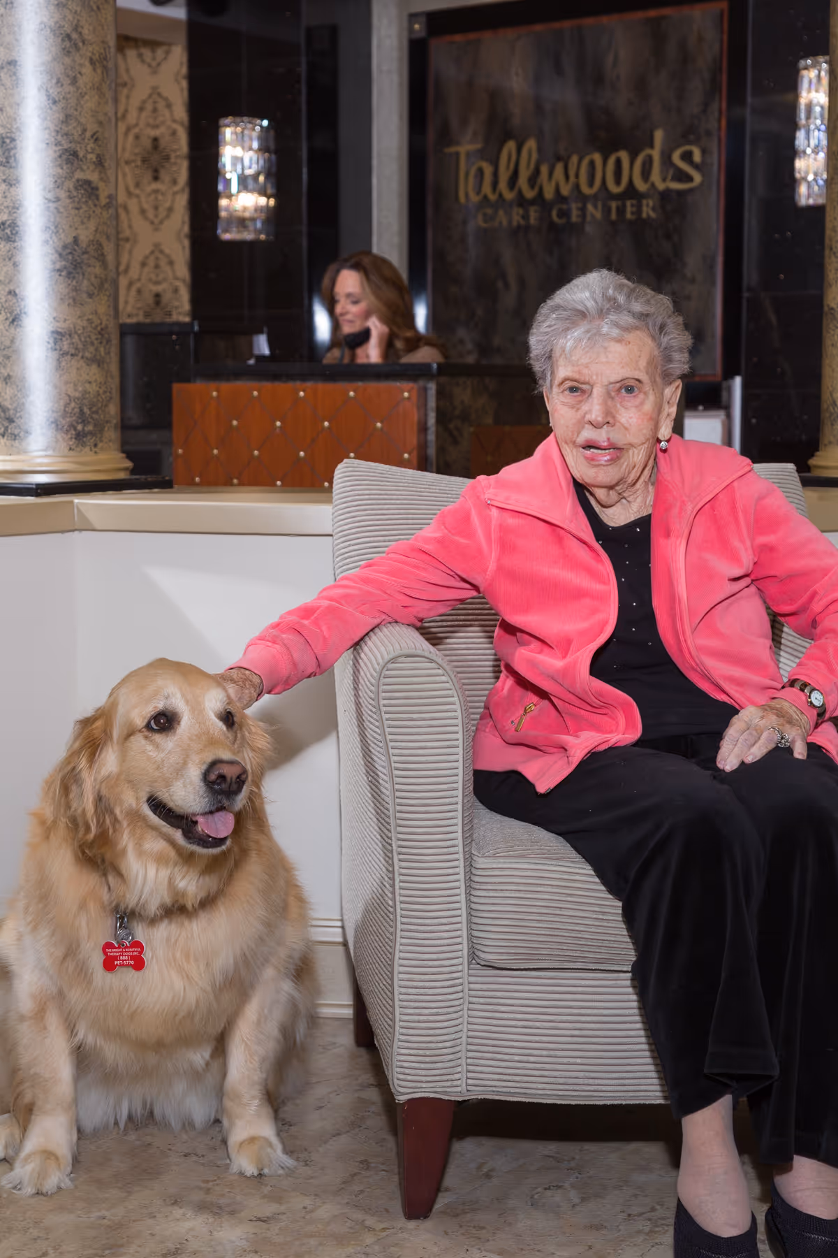 An elderly woman wearing a pink jacket sits in a beige armchair inside Tallwoods Care Center, petting a golden retriever sitting beside her. In the background, a receptionist is visible behind a counter with the Tallwoods Care Center sign on the wall.