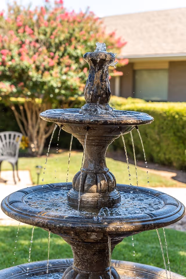 A close-up view of a three-tiered stone water fountain with water flowing from the top tier down to the lower basins. In the background, there is a green lawn, a tree with pink flowers, a hedge, a building with a window, and a white outdoor chair.