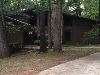 A rustic, single-story building with dark wooden exterior walls surrounded by tall trees and greenery. A concrete pathway leads up to the building, which has large windows and a sloped roof.