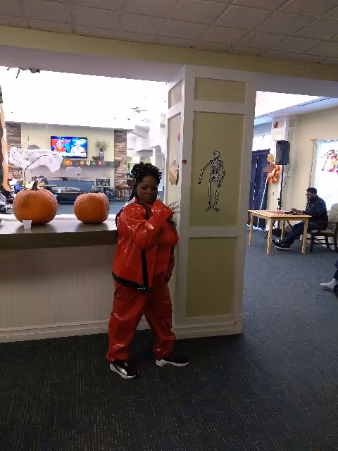 A person dressed in a red outfit posing indoors near a counter with two pumpkins on it. The room has Halloween decorations including a skeleton drawing on a pillar and a ghost decoration. In the background, there is a television mounted above a fireplace and a person sitting at a table.