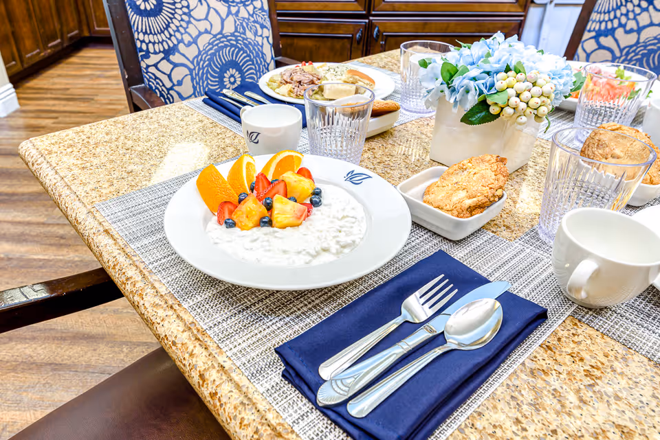 A dining table set with plates of food including cottage cheese with fruit, a sandwich, cookies, and glasses of water. The table has a beige granite surface with a gray woven placemat, silverware on a navy blue napkin, and a small floral centerpiece. Blue patterned chairs and wooden cabinetry are visible in the background.