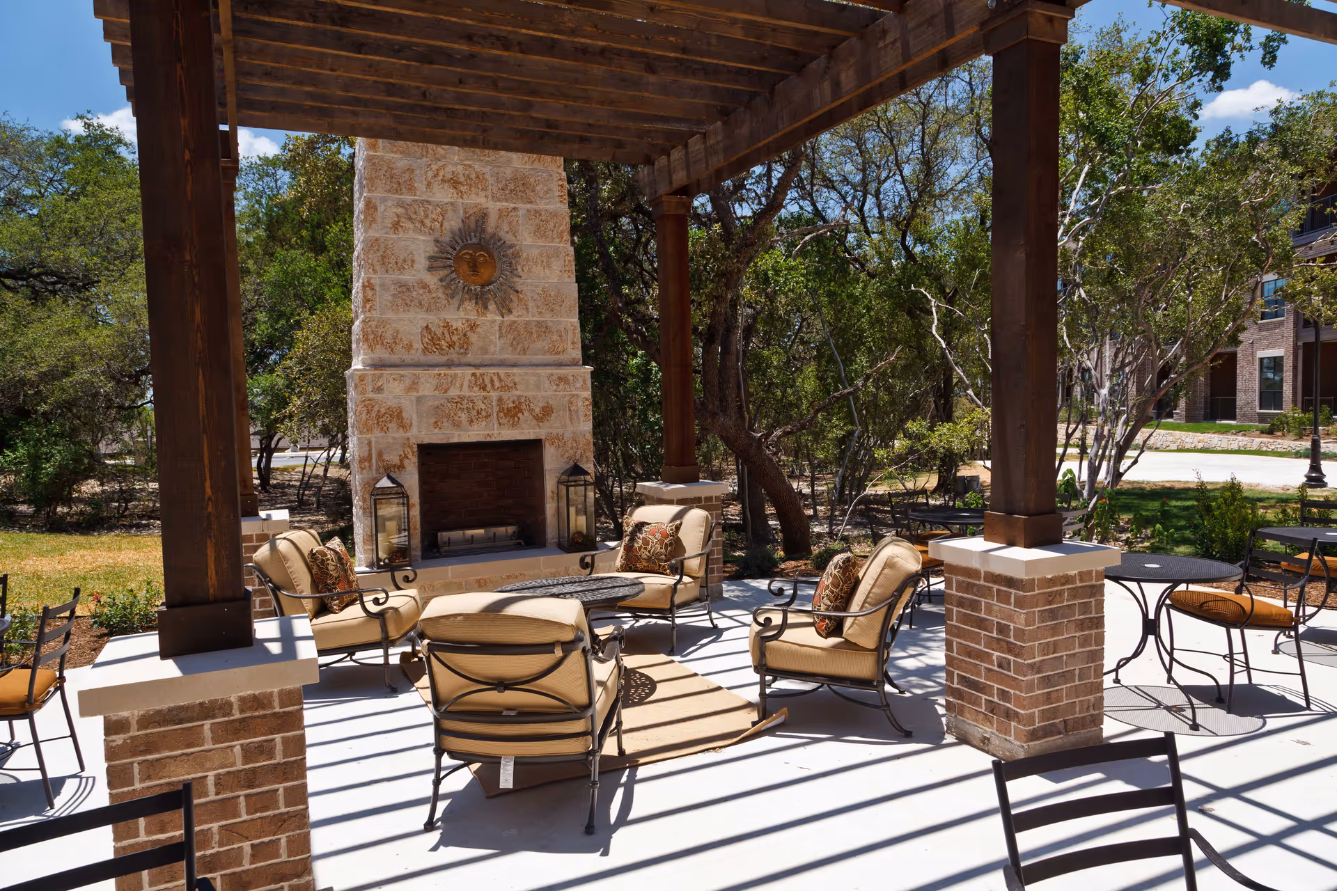 Outdoor seating area with cushioned chairs arranged around a stone fireplace under a wooden pergola. The area is surrounded by trees and greenery, with additional tables and chairs nearby on a sunny day.