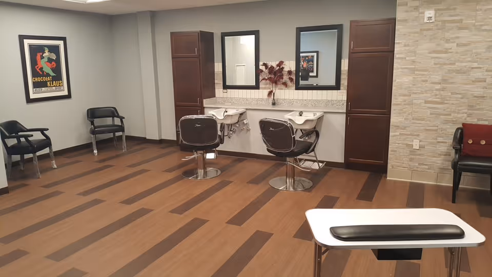 Interior view of a salon area with two black salon chairs in front of two sinks and mirrors, wooden cabinets on either side, three black waiting chairs along the walls, a framed poster on the wall, and a manicure table in the foreground.