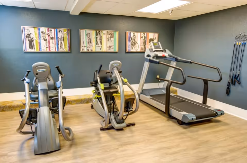A small fitness room with two recumbent exercise bikes and a treadmill on a light wood floor. The walls are painted dark gray and decorated with three framed vintage-style black and white photos of people exercising, each bordered with colorful stripes. On the right wall, several resistance bands are hanging on hooks.