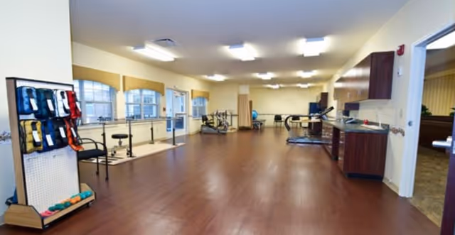 Spacious bright rehabilitation/fitness room with wood floors, parallel bars, exercise equipment, and cabinets.