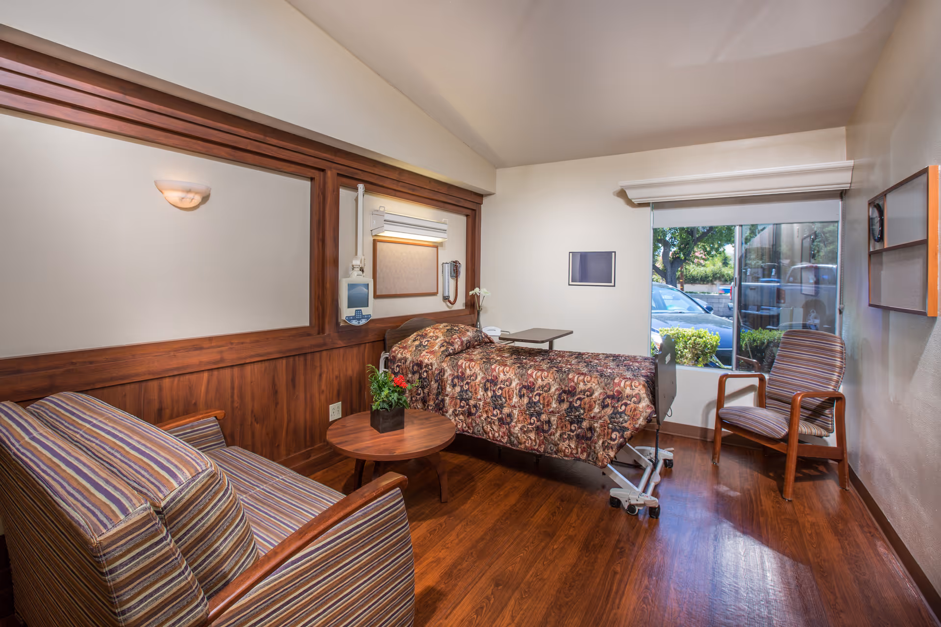 A cozy room in Woodland Care Center featuring a single hospital-style bed with a patterned bedspread, a small round wooden table with a plant, a striped armchair, and a window showing a view of parked cars and greenery outside. The room has wooden flooring and wood paneling on the walls.