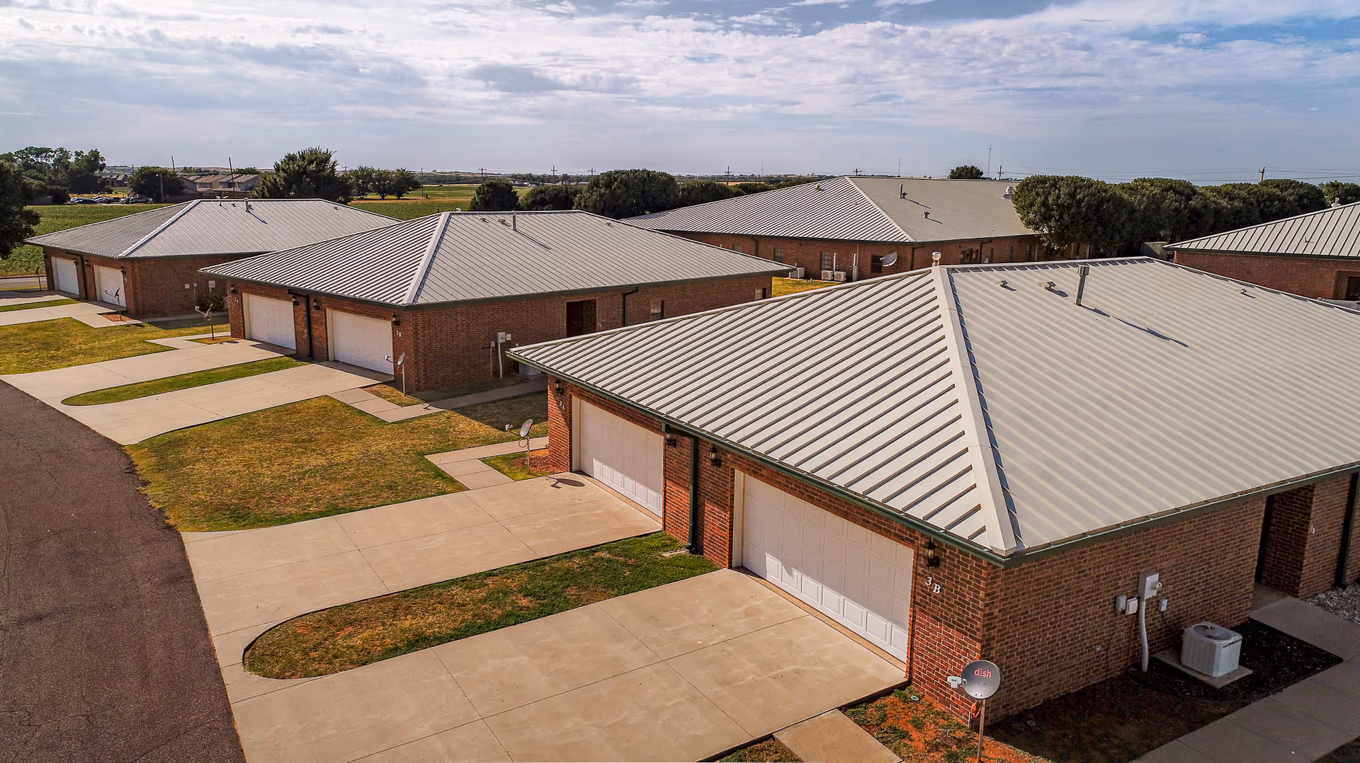 Aerial view of multiple single-story brick buildings with metal roofs, each featuring a driveway and garage, set in a senior living community with green lawns and a partly cloudy sky.
