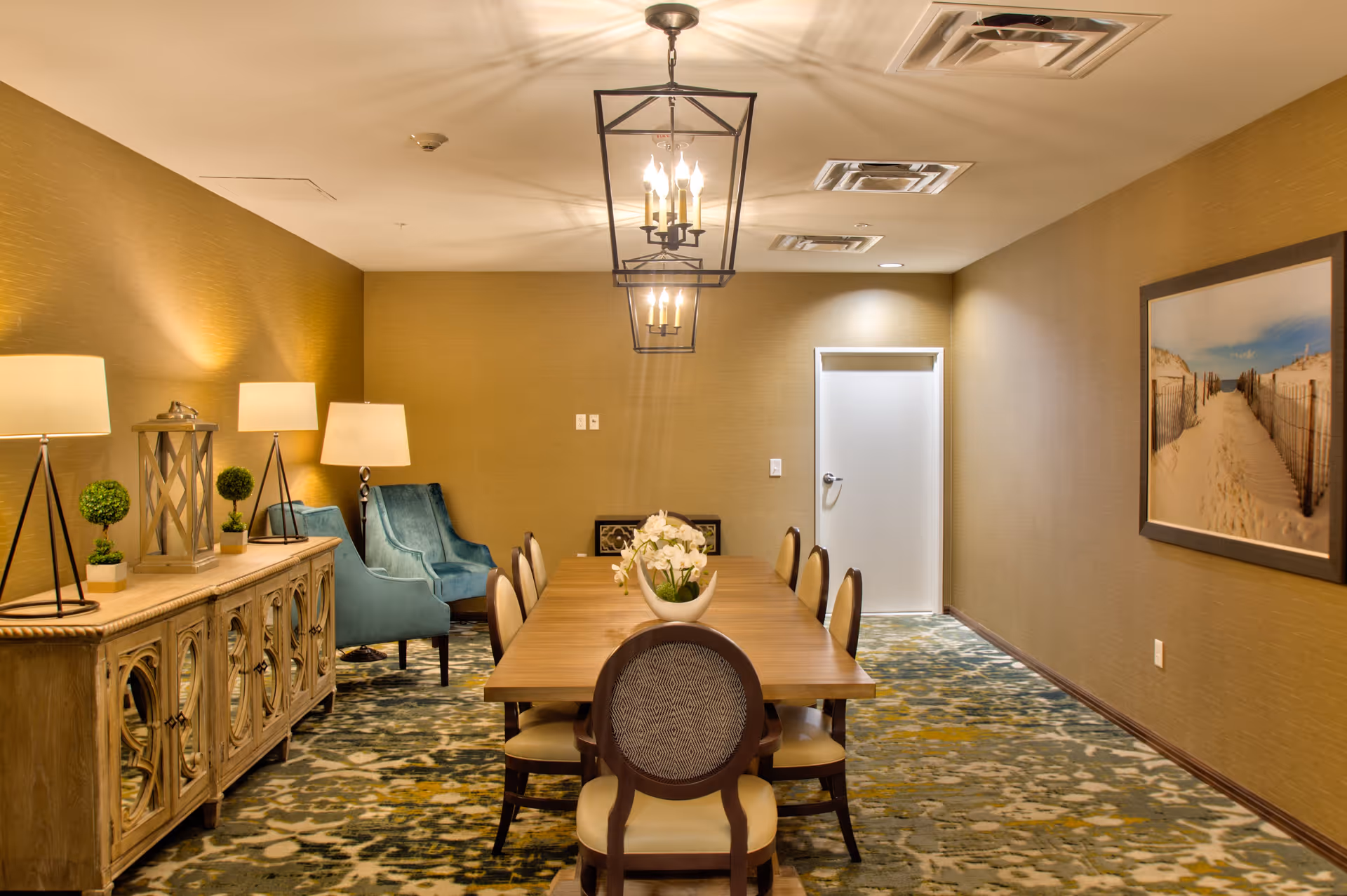 A well-lit dining room with a long wooden table surrounded by eight chairs. The room features warm beige walls, a patterned carpet, two blue armchairs in the corner, and a sideboard with decorative lamps and plants. A framed picture of a sandy beach pathway hangs on the right wall.
