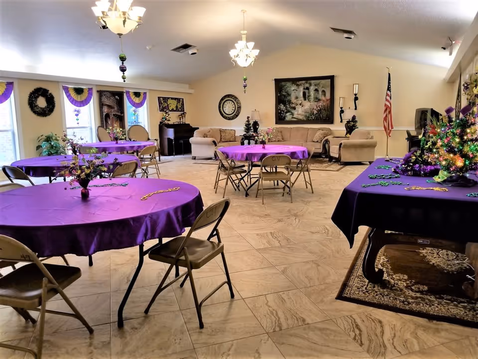 A spacious common area decorated with purple tablecloths on round tables, folding chairs, and festive Mardi Gras decorations including beads and wreaths. The room features beige tiled flooring, beige walls, chandeliers, a beige sofa set against the far wall, a piano, a large wall clock, a framed painting, and an American flag in the corner.