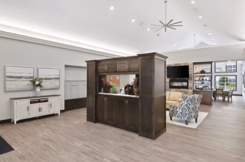 A spacious and well-lit senior living facility interior featuring a large dark wooden aquarium divider in the center. To the right, there is a cozy seating area with a beige sofa, a patterned armchair, a wall-mounted TV above a stone fireplace, and large windows letting in natural light. On the left side, there is a white cabinet with two framed abstract paintings above it and a vase with flowers. The floor is light wood, and the ceiling is vaulted with recessed lighting and a large ceiling fan.