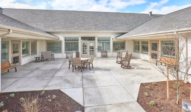 Outdoor courtyard area with concrete flooring surrounded by a single-story building. The courtyard features several wooden benches, rocking chairs, and a table with chairs. There are small garden beds with some plants and young trees around the edges. The sky is partly cloudy.