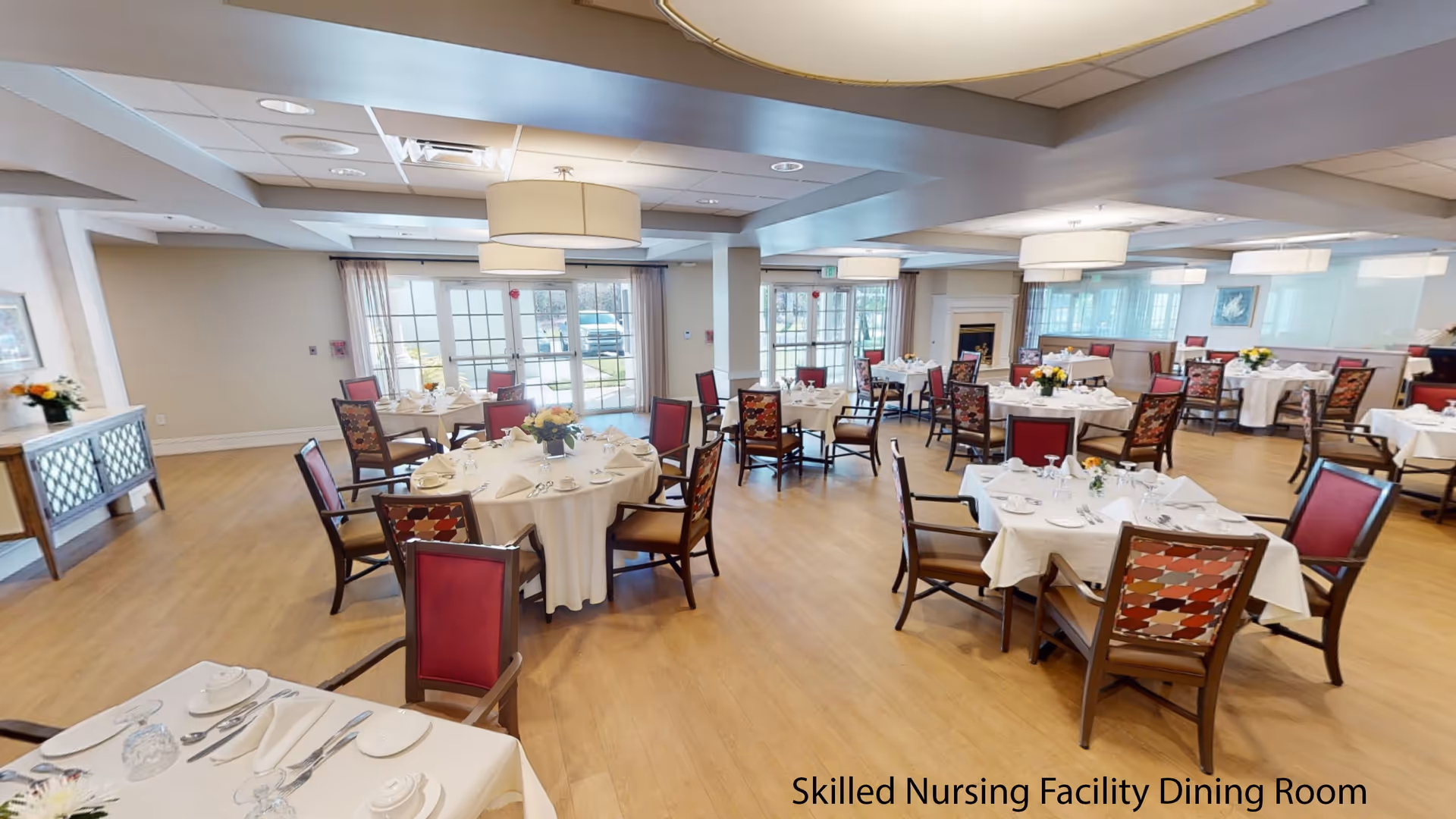 A spacious dining room in a skilled nursing facility with multiple round and square tables covered with white tablecloths, set with plates, cups, napkins, and silverware. The chairs have red and patterned upholstery. Large windows and glass doors allow natural light to fill the room. Modern ceiling lights hang above the tables, and there is a fireplace in the background.