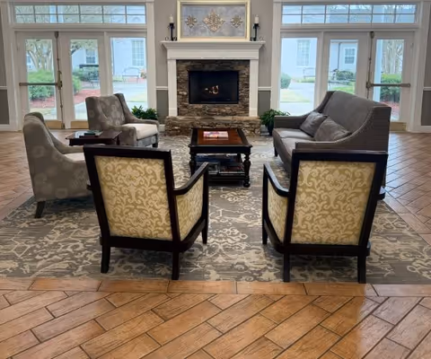 A cozy living room area with a stone fireplace in the center, flanked by two large windows on each side. The room features a patterned area rug, two armchairs with yellow and beige upholstery, two gray armchairs, and a gray sofa. A wooden coffee table sits in the middle with books on it. The floor is tiled with a wood-like pattern.