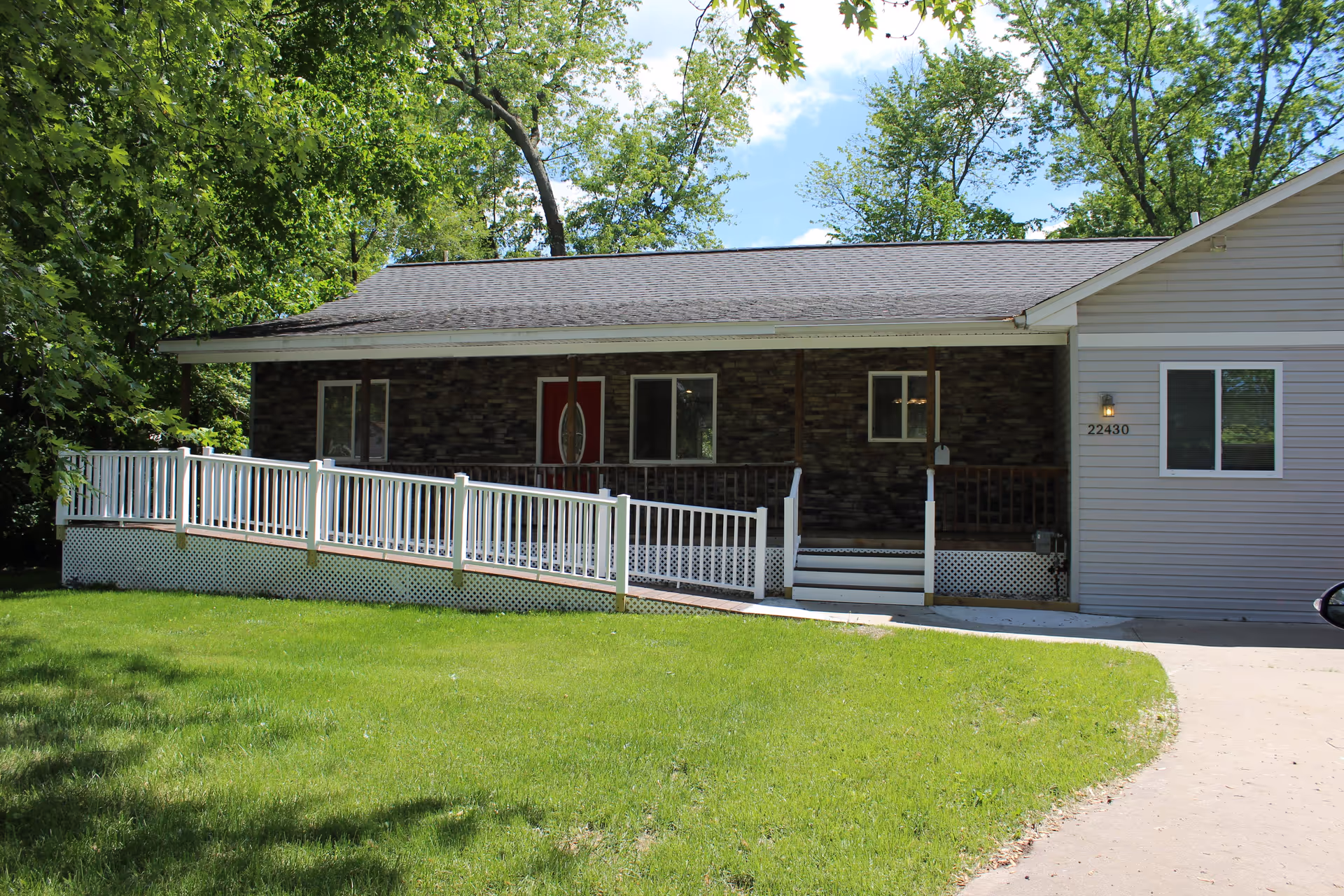 Single-story residential building with a front porch featuring a white ramp and stairs. The building has a stone facade section with a red front door and windows, and a light gray siding section with a window and the number 22430. The area is surrounded by green grass and trees under a blue sky with some clouds.