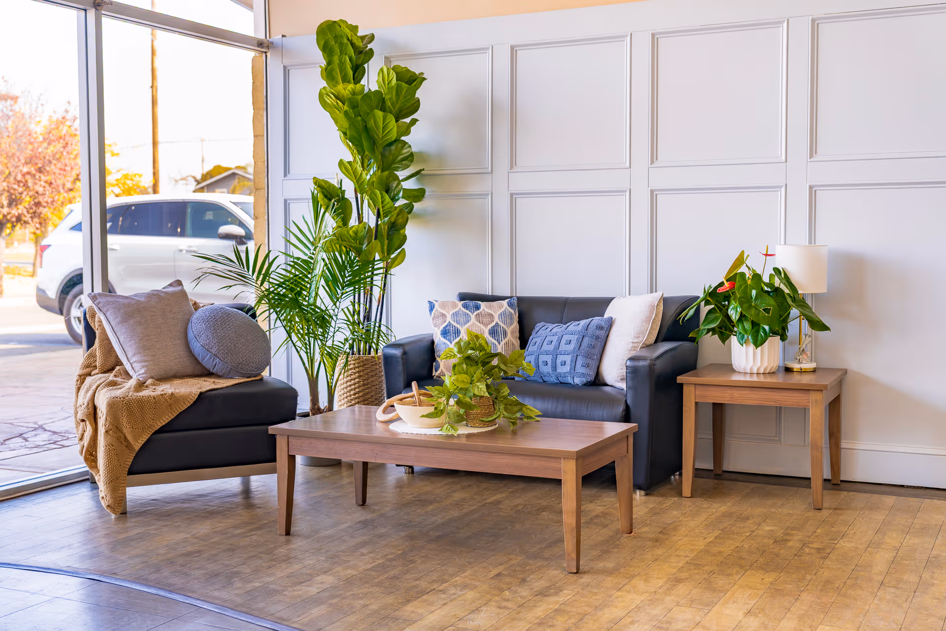 Bright seating area with a sofa, armchair, wooden coffee and side tables, and several potted plants by large windows.